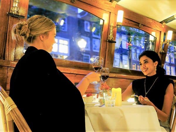 Two friends laughing and toasting with wine on a dinner cruise in Amsterdam.